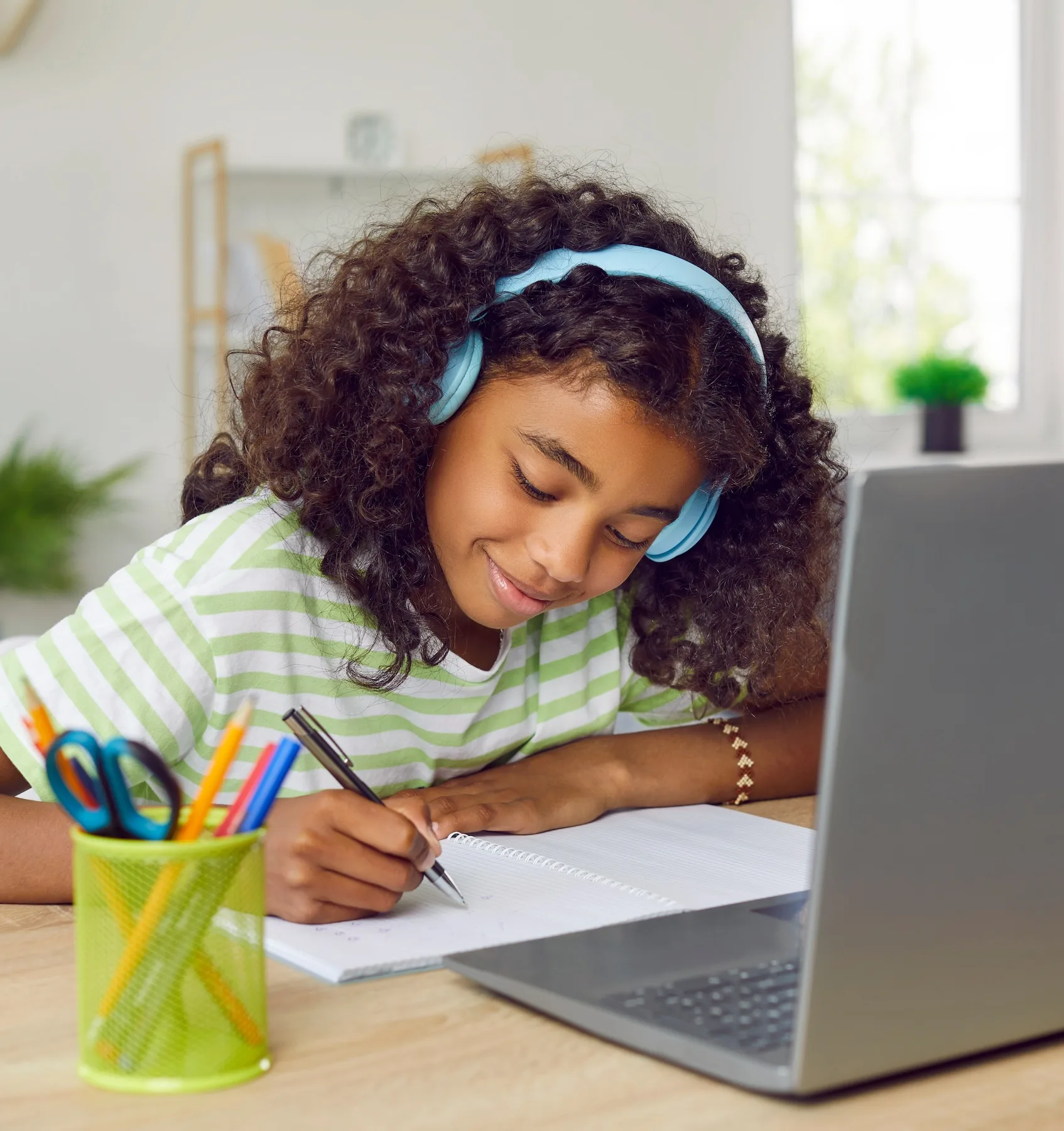 A girl wearing blue headphones and sitting in front of a laptop, drawing on a notepad.