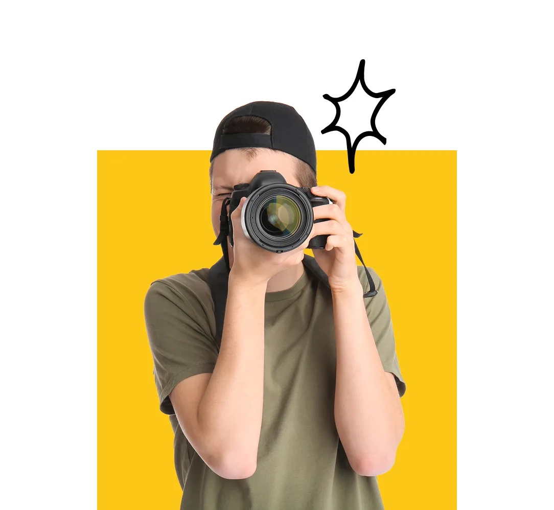 A young man in a green shirt aiming a camera at the viewer with a solid yellow square background